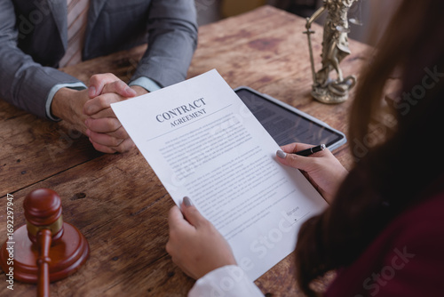 Two lawyers are sitting at a wooden table reviewing a contract agreement, with a gavel and statue of Themis nearby, symbolizing justice and legality
