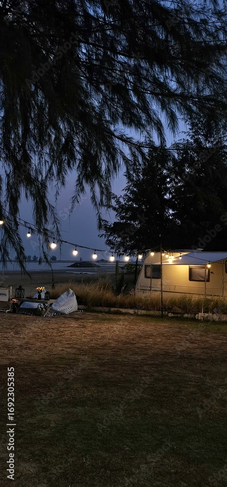 Obraz premium Beach Campsite with a Caravan and Lights.A vertical shot of a cozy campsite scene on a beach at dusk. A white caravan is parked on the dark, grassy ground, with a string of warm, glowing lights hangin