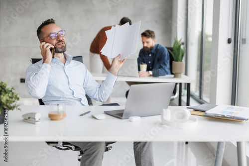 Busy professional in bright office space organizing documents while talking on phone during work hours