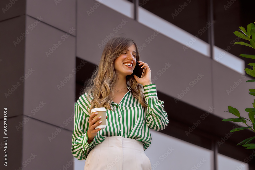 Fototapeta premium Professional Hispanic Woman Talking on Phone and Holding Coffee Outdoors in Modern Setting. Business person portrait on a bright daylight