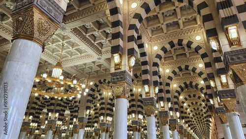 The image showcases the interior of Al-Masjid an-Nabawi in Medina, Saudi Arabia. It features a series of grand arches with alternating black and white stone patterns, supported by sleek marble columns