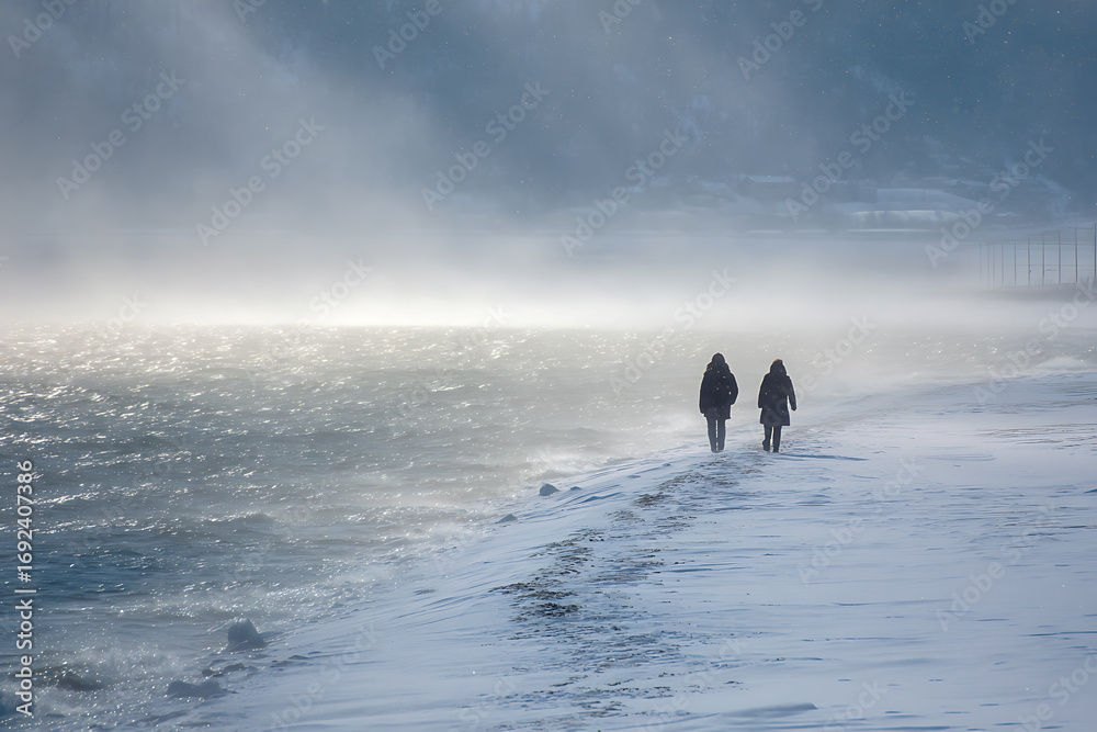 Fototapeta premium Winter walk along the snowy beach with dramatic light and two figures silhouetted against the misty horizon.