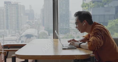 Senior Asian businessman wearing headset celebrates success during online meeting in modern co-working space. Remote work and business communication concept	
