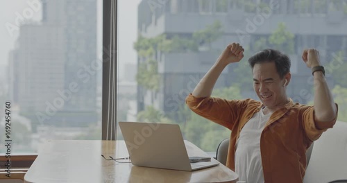 Senior Asian businessman wearing headset celebrates success during online meeting in modern co-working space. Remote work and business communication concept	
