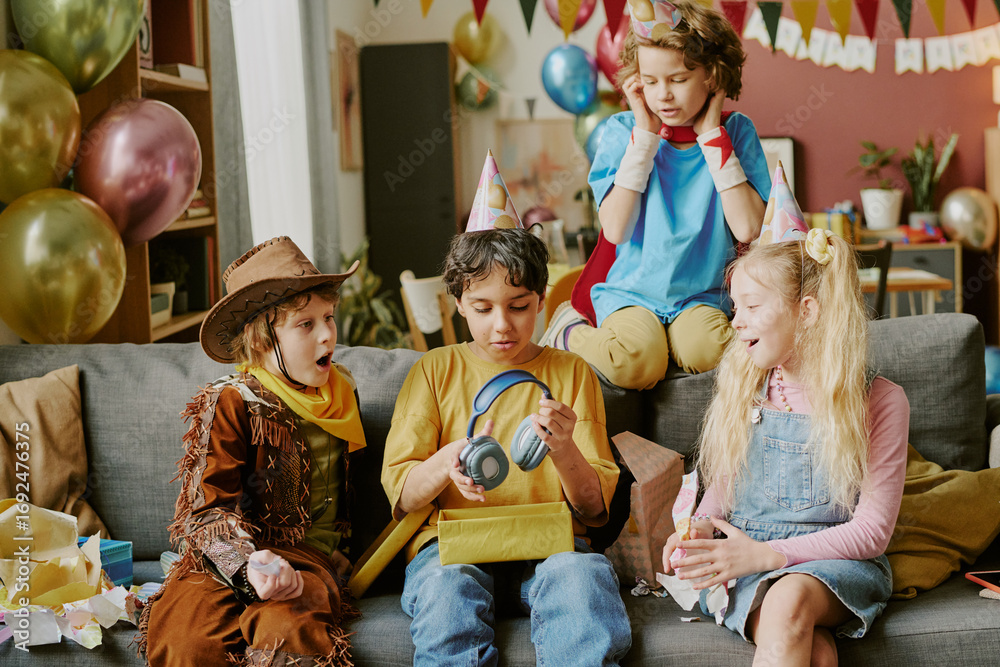 Obraz premium Group of four children including two girls and two boys of diverse ethnicities sitting on sofa wearing party hats, opening presents and celebrating birthday with decorations and balloons