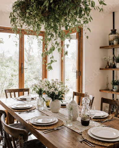 Natural bohemian dining room with wooden table, white ceramics, woven placemats, greenery, and sunlight window view