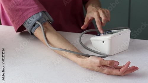 Young woman in pink t-shirt measures blood pressure with tonometer sitting at table. Concept of healthy heart and self-care