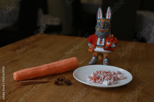 A decorative rabbit figurine with carrots, spices, and a sweet treat on a wooden table.