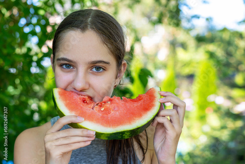Teen girl eating juicy watermelon outdoors. Portrait of a teenage girl enjoying a slice of fresh watermelon in a green garden setting during a warm summer day.