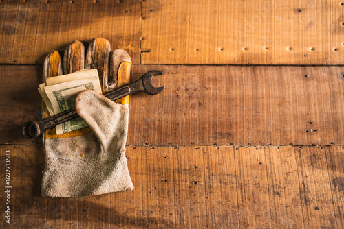 Leather work gloves holding a rusty wrench and American dollars on a rustic wooden work table