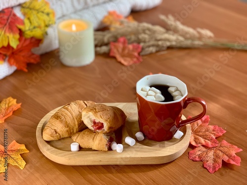 A cozy setting features a cup of coffee topped with marshmallows alongside croissants filled with strawberries. Autumn leaves decorate the wooden table, enhancing the seasonal ambiance