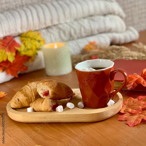 A cozy setup featuring a cup of hot coffee alongside croissants and marshmallows on a wooden tray. Autumn leaves add a seasonal touch to the inviting atmosphere