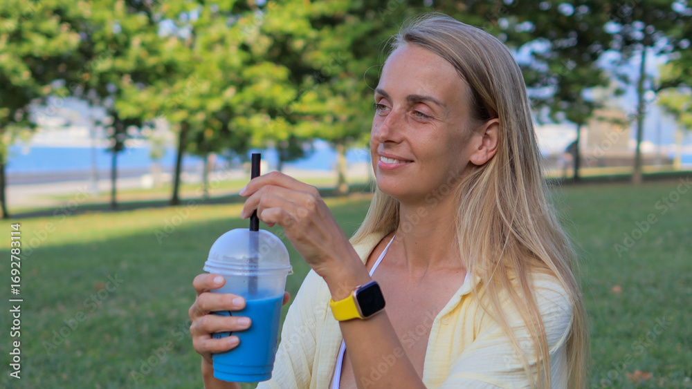 Fototapeta premium Woman relaxing in park and drinking blue matcha tea with straw