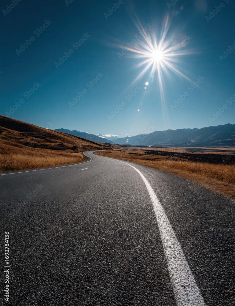 Naklejka premium A winding asphalt road stretches into a mountainous landscape under a vibrant blue sky, highlighted by a sunburst