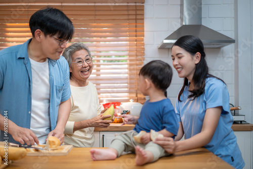 Asian family spending quality time in the kitchen — a grandmother playfully bites into a mango while surrounded by her son, daughter-in-law, and grandson during food preparation.