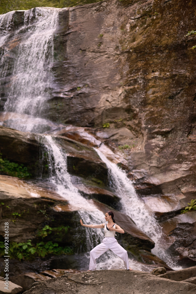 Naklejka premium woman performing slow tai chi movements near a waterfall and river at sunset capturing harmony balance and fitness in nature