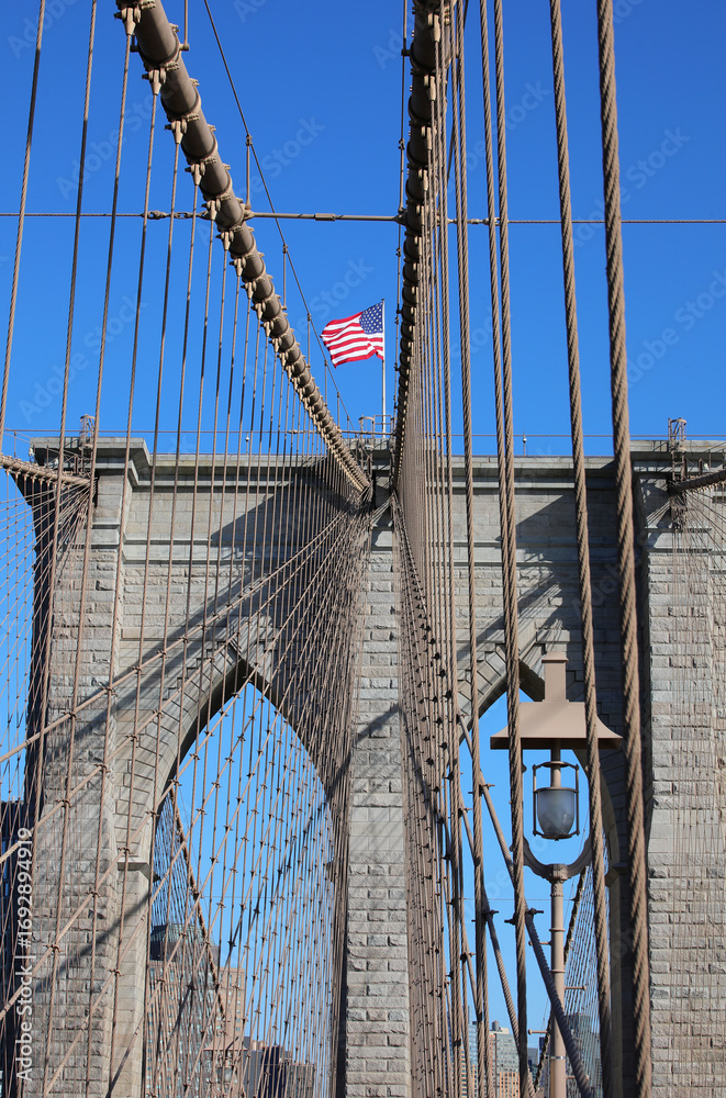 Fototapeta premium New York city with Waving American Flag on the top of Tower of Brooklyn Bridge and steel cables