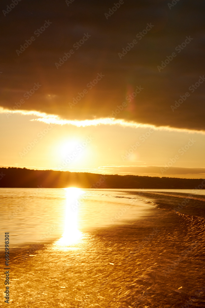 Obraz premium Golden sunset over river with dramatic clouds and bright reflections on water. Autumn trees glowing in warm evening light, creating a peaceful natural landscape with rippling waves