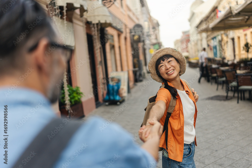 Obraz premium Tourist woman leading her partner by hand while walking through a city street lined with cafes and shops