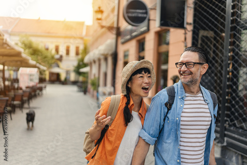 Fototapet Smiling tourists in their forties holding hands and walking through a charming c
