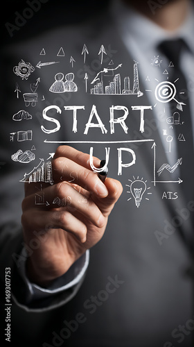 Closeup of a mans hand in a grey suit writing Startup with a black stylus on a dark background surrounded by white sketched business icons
