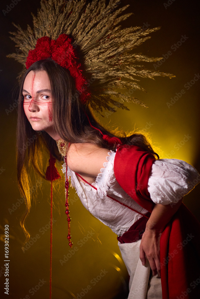 Naklejka premium Mystical woman with striking red face paint and traditional headdress stands against a golden, dramatic background, exuding an air of ancient power and enigmatic beauty .