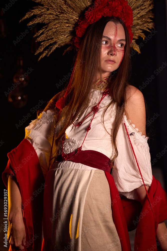 Naklejka premium Portrait of a young woman in traditional Ukrainian costume with red embroidery, a headpiece of dried wheat and red pom poms, and red face paint looks off to the side at a dark background .