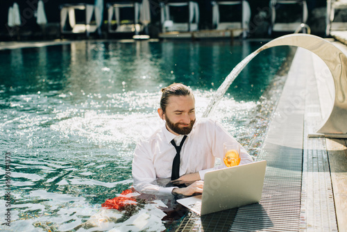 Businessman working on laptop in swimming pool with cocktail