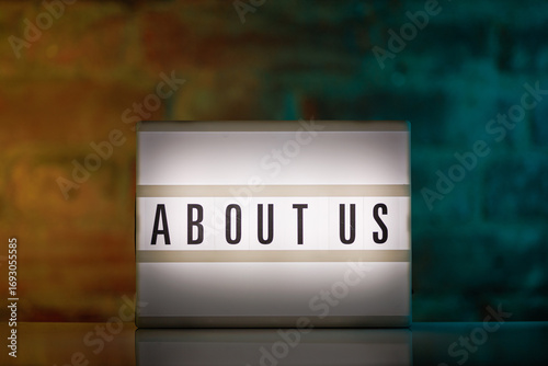 Photos A cinematic lightbox with the words About Us illuminated, placed against a rusti