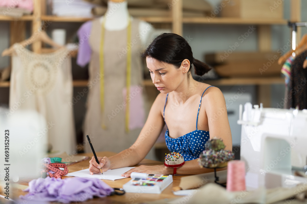 Fototapeta premium Young woman dressmaker draws sketch of clothing on paper in workshop