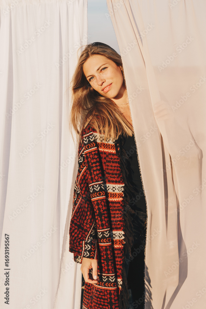 Fototapeta premium Beautiful young woman is looking to the camera, photo shoot with textile background on the meadow. End of summer, girl is happy and enjoying warm sunny day. Author color correction and stylish outfit 