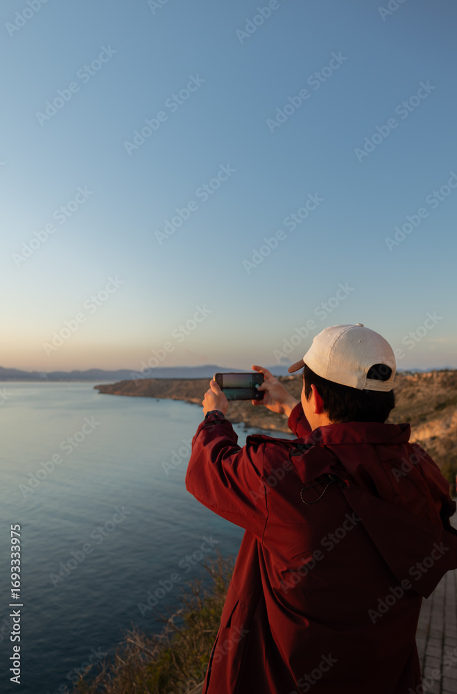 Obraz premium Young man taking a photo with his cell phone of the seascape at an incredible sunset, concept of tourism, technology, inspiration, and lifestyle