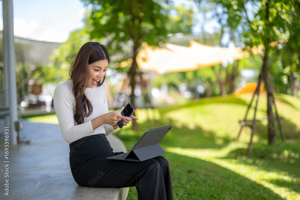 Fototapeta premium Excited businesswoman comparing smartphone and tablet in park