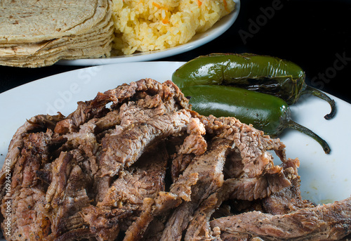 Charcoal-grilled beef with grilled jalapeño peppers on a white plate to make tacos as Mexican food with tortillas and rice on the bottom.