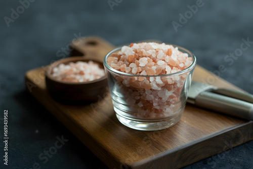 Himalayan Pink Salt Presentation: A close-up shot of Himalayan pink salt crystals in a glass and wooden bowl. The crystals sit atop a wooden cutting board.  A metal spoon is beside the glass.