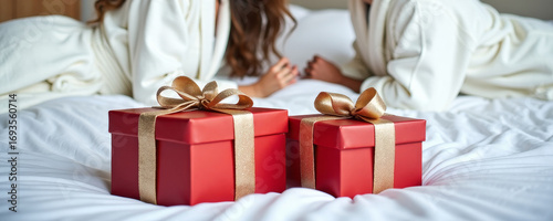 Red Christmas gifts boxes wrapped in red paper lying on white bed in foreground, creating cozy holiday atmosphere. Couple of man and woman on background.