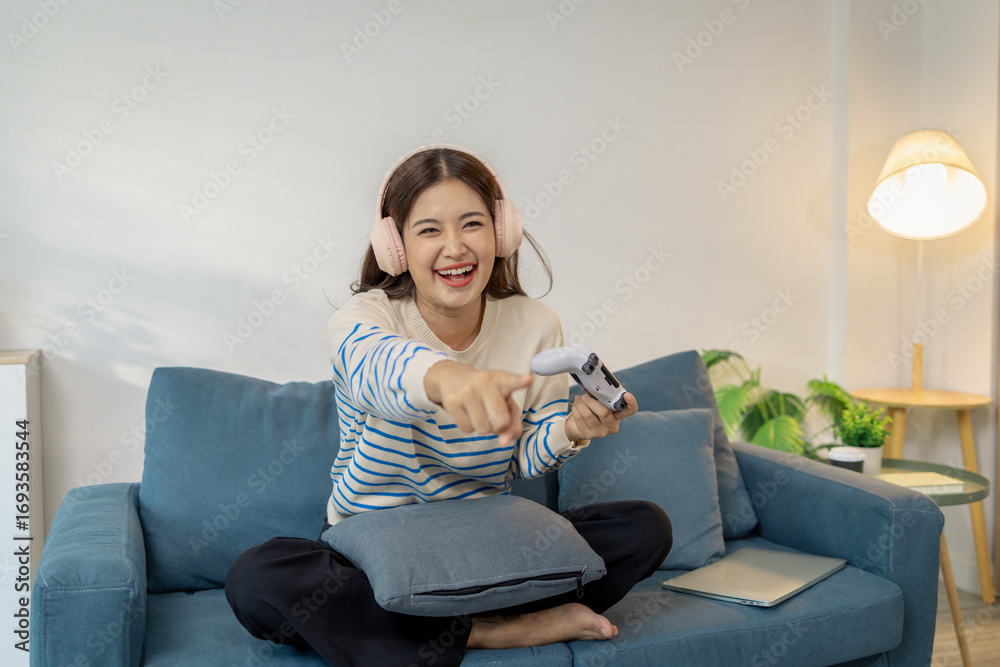 Obraz premium A young woman wearing headphones is sitting on a sofa while holding a game controller, showing joy, excitement, and focus as she plays a video game indoors.