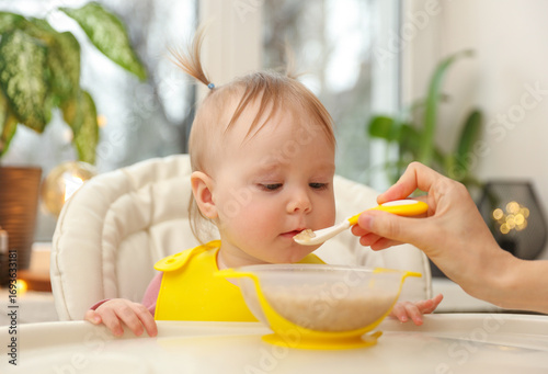 Mother's hand feeds her cute little baby daughter at the feeding table