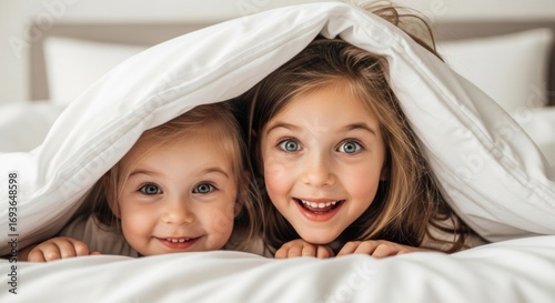Two young girls peek out from under a white duvet, smiling and looking directly at the camera with bright, joyful expressions.
