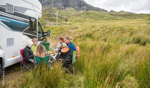 Family group sharing a meal outdoors next to a camper van in the Scottish Highlands surrounded by mountains and grassy landscape