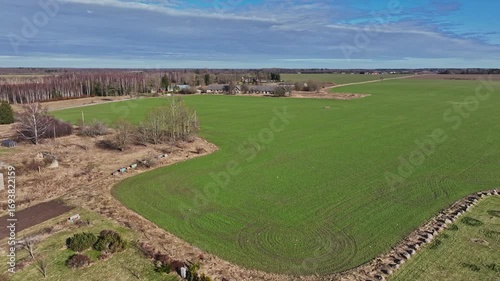 Forward-moving drone footage of green farmland in Paide, Estonia. Freshly sprouted field with tractor tire patterns, bordered by dry grass, scattered trees, rocks and small sheds near the edge.