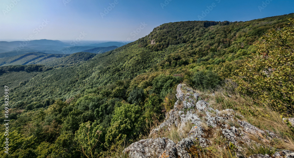 Obraz premium Scenic view of the rocky ridge at Három-kő saddle in the Bükk Mountains, Hungary. The rugged cliff contrasts with the lush green forested slopes.