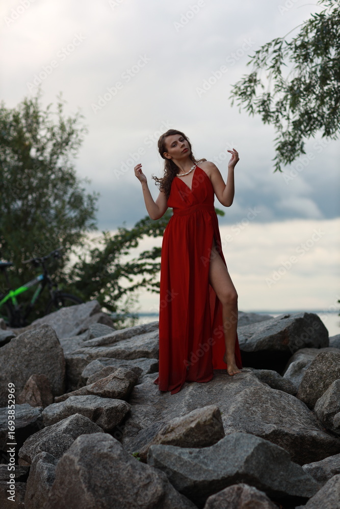 Fototapeta premium Pretty slim brunette woman in red dress standing full length on the rocks near green bushes