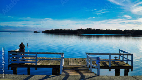 The entrance NSW Australia view of the ocean and bay.People on the beach in the evening.Fishing from a wooden dock on a serene lake at sunrise with calm water reflecting the blue sky