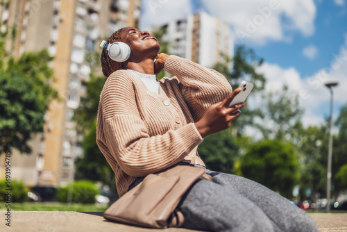Portrait of a cheerful black woman in her 20s using her mobile phone outdoors in an urban setting, wearing wireless headphones and smiling as she texts friends, embodying modern city life and leisure.