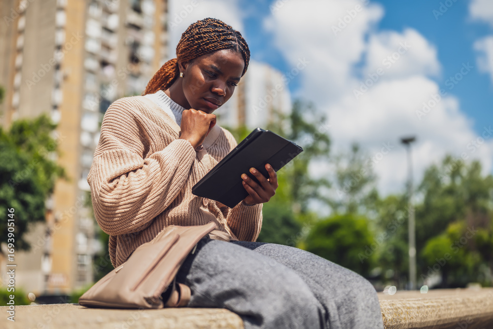 Fototapeta premium African American woman in her 20s looks worried and displeased as she interacts with her tablet. She is seated outdoors reflecting the pressures of city life.