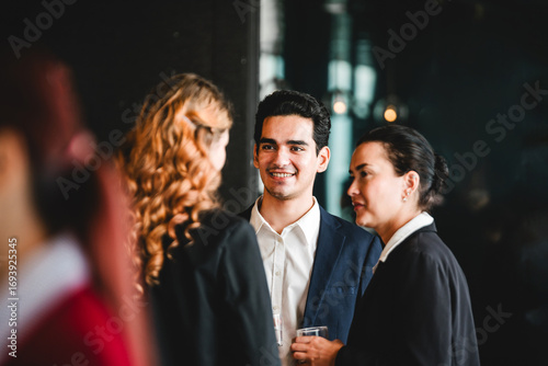 Professionals in formal attire converse and smile at indoor corporate event. Business networking scene includes meeting, conference, seminar, teamwork, discussion, colleagues, and collaboration.