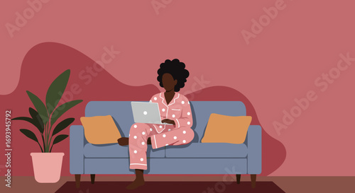 African American woman in pajamas works on her laptop from a comfortable home sofa, illustrating remote work, relaxation, and modern lifestyle.