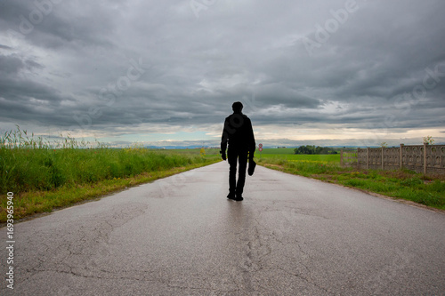 silhouette di uomo che cammina, in viaggio su strada, ripresa dal basso, che si perde all'infinito tra erba alta verso il cielo denso di nuvole.
