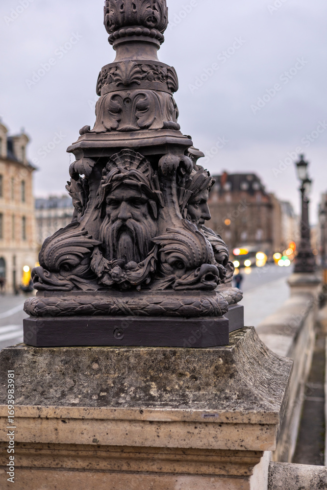 Fototapeta premium Close-up of an ornate, historic lamppost on the Pont Neuf bridge in Paris, France. The cast iron base features grotesque mascarons and sea creatures.